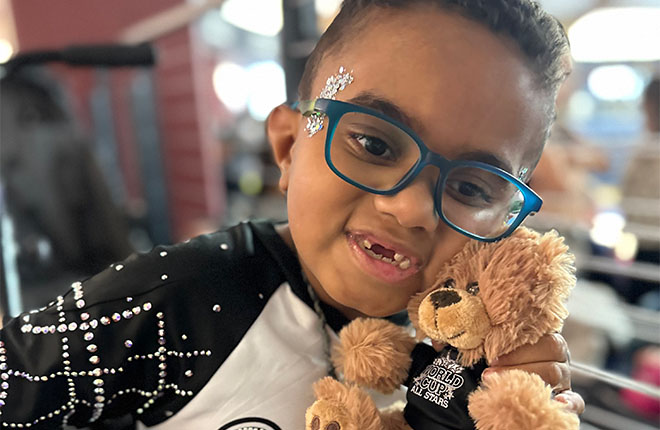 Mikey Stipeck in a white shirt holding a teddy bear, indoors with festive lights in the background.