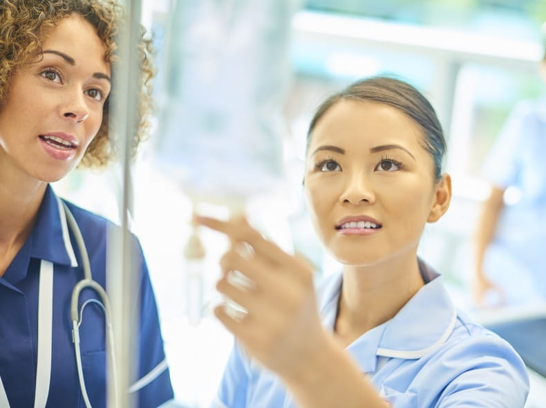 A senior nurse is supervising a young nurse as she attends to a saline drip on a hospital ward.