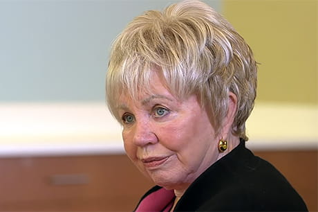 An older woman with short, blond hair and wearing gold earrings sits indoors, facing slightly to the left with a neutral expression.