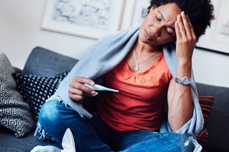 Young African American woman holding her head and taking her temperature while sitting on a couch.