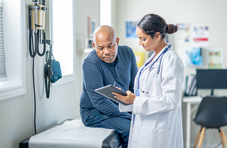 A senior gentleman sits up on an exam table. A female doctor is holding out a tablet as they review test results together.
