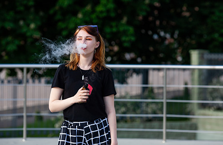 A female teen vapes while standing outside.