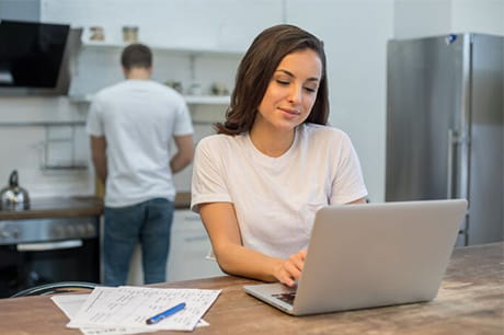 Beautiful young woman looking at gadget screen while her husband cooking on the background at the kitchen. Relationships concept